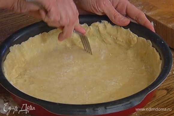 На дно круглой формы выложить круг из бумаги для выпечки и равномерно распределить тесто, сформировав бортики, затем часто наколоть его вилкой.