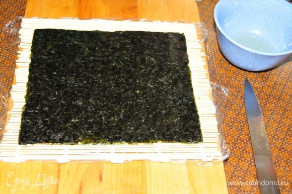 Начинаем делать роллы. Оборачиваем коврик для приготовления роллов пищевой плёнкой. Кладём его на стол. Сверху укладываем лист сушёных водорослей – нори.
