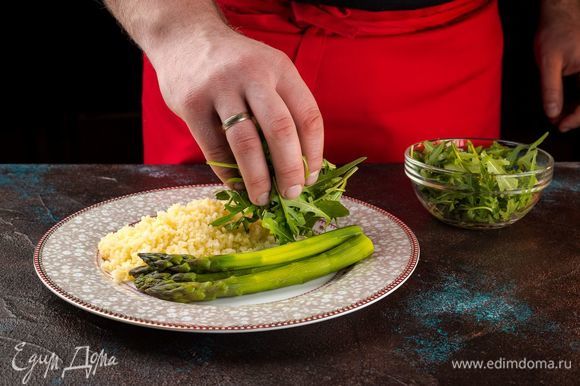 Выложите кускус со спаржей порционно в тарелки, сверху положите руколу и полейте блюдо оливковым маслом.
