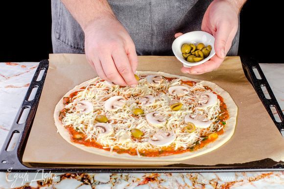 Сверху на пиццу выложите натертый сыр, нарезанные шампиньоны и разрезанные пополам оливки.