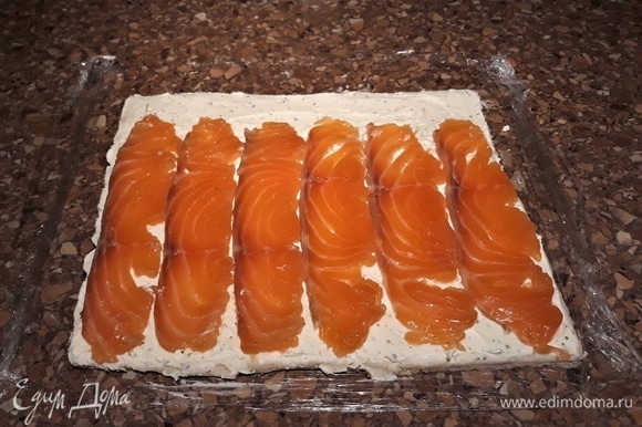 На пищевую пленку уложите хлеб слегка внахлест по три кусочка в два ряда. Скалкой примните, чтобы не было швов. Смажьте хлебный пласт творожно-сливочным сыром. Сверху выложите полоски рыбы.