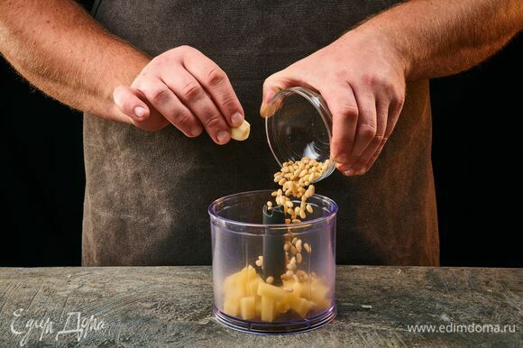 Чтобы приготовить песто, выложите в чашу блендера пармезан, зубчик чеснока и кедровые орехи.