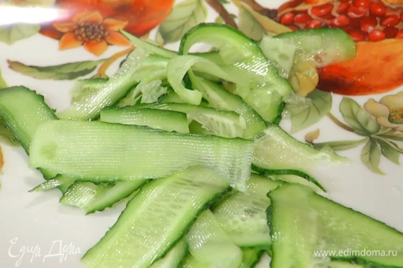 Огурец с помощью овощечистки нарезать тонкими полосками и выложить в большую тарелку, посыпать измельченным луком и листьями тархуна.